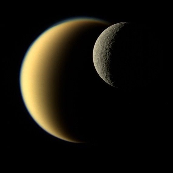 Titan as a hazy gold crescent with the cratered moon Rhea passing in front